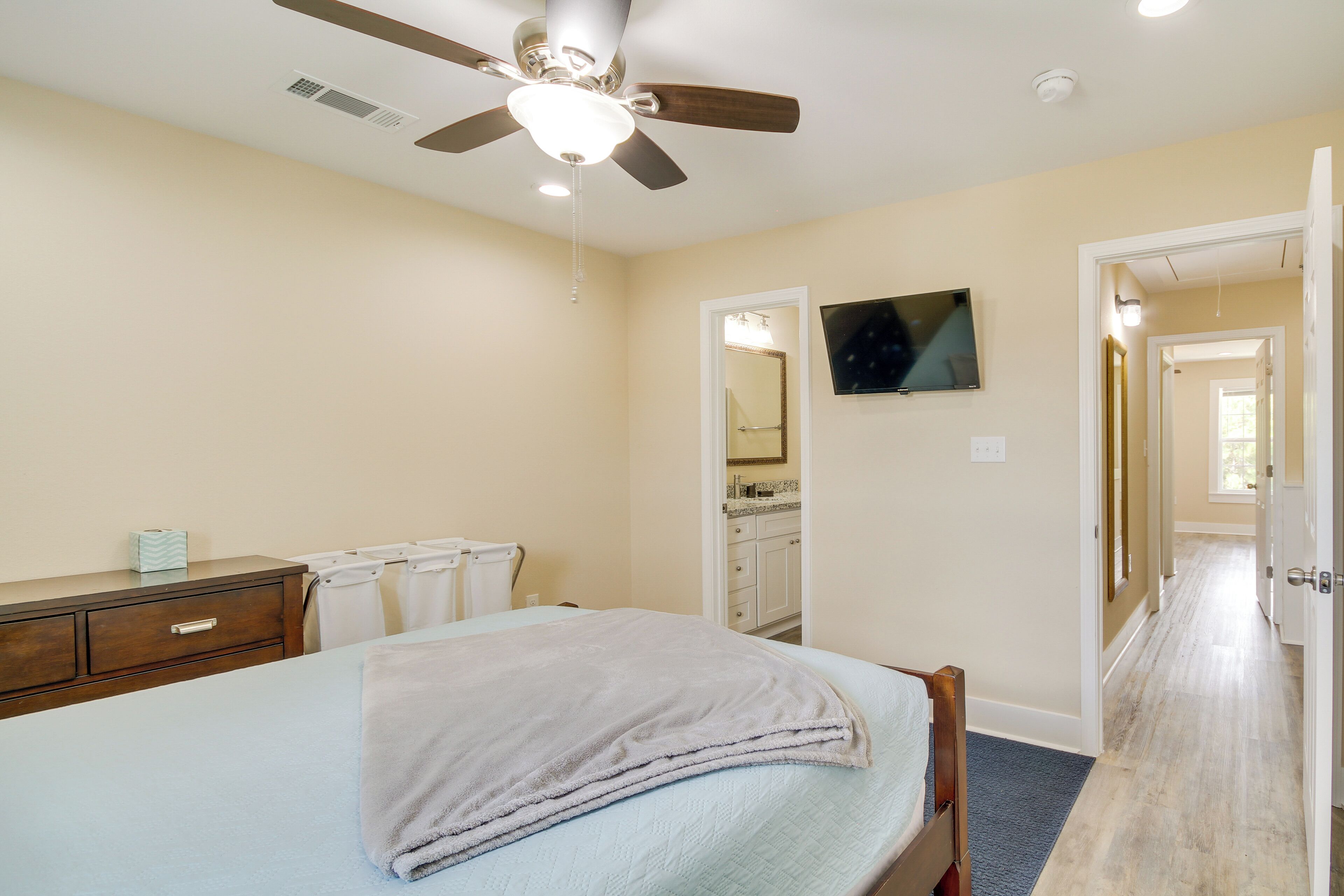 Une chambre à coucher avec un lit, un ventilateur de plafond, une télévision, une commode et une salle de bain visible à travers une porte ouverte.