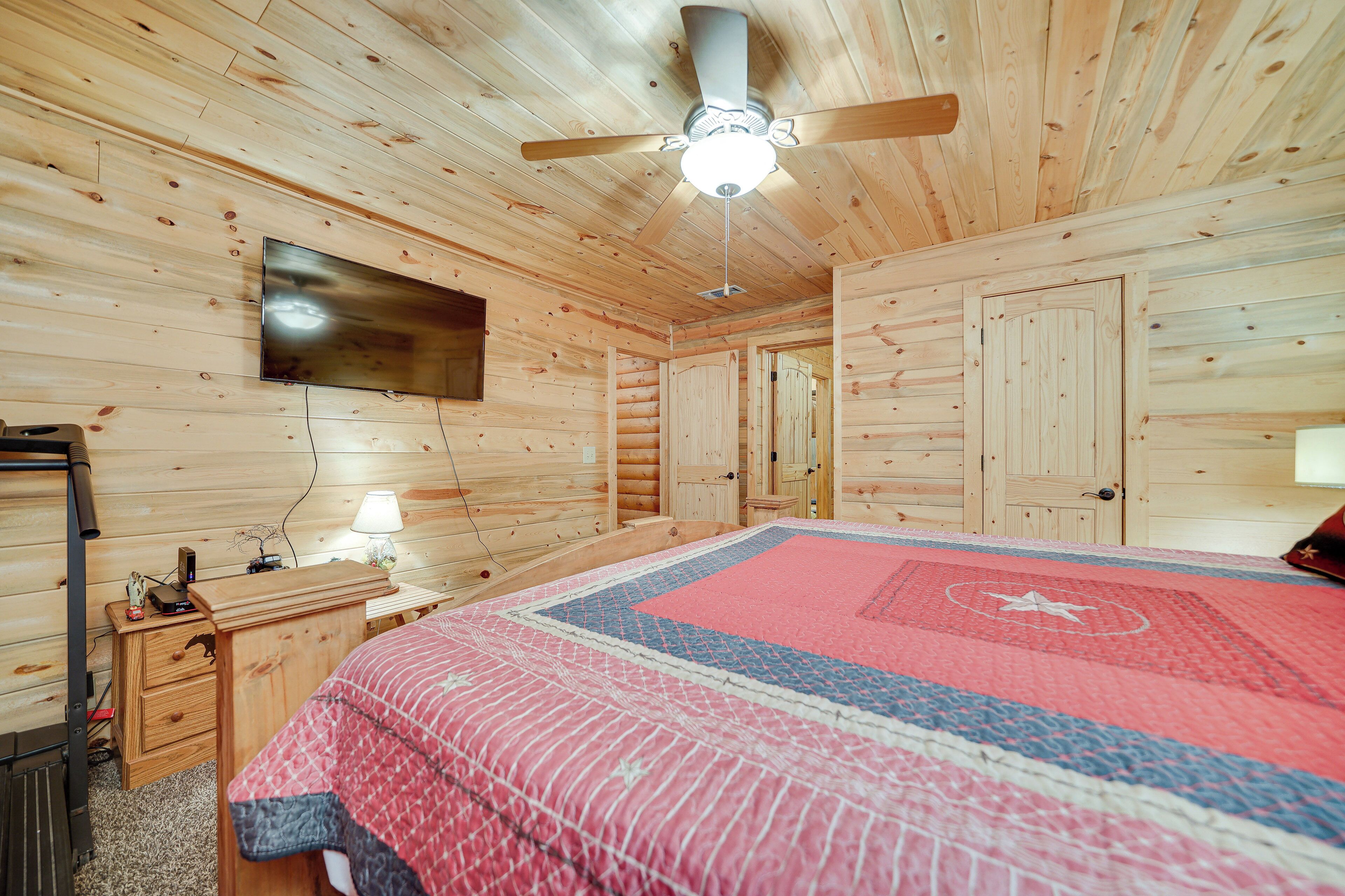 A wooden cabin bedroom with a bed, a ceiling fan, a TV mounted on the wall, and a wooden dresser.
