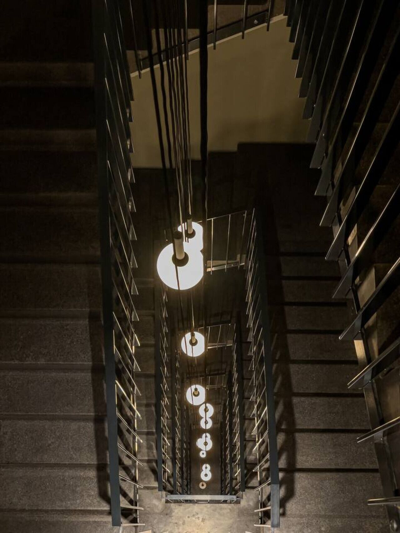 Ein modernes Interieur mit einer zentralen Treppe, begleitet von Metallgeländern und hängenden Leuchten.