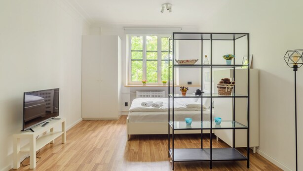 Une chambre moderne avec un lit, un téléviseur posé sur un meuble, une fenêtre donnant sur un paysage verdoyant et une étagère en métal noir agrémentée d’objets décoratifs.