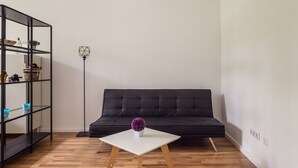 Un salon moderne avec un canapé noir, une table basse blanche ornée d’une fleur violette et une étagère métallique noire adossée à un mur blanc.