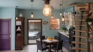 Une cuisine moderne, dotée d’un éclairage suspendu, d’une table à manger avec des chaises, et offrant une vue sur une voiture à travers une porte vitrée.