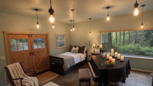 Un dormitorio con cama, piano, mesa de comedor con velas, sillón mecedora y una ventana grande con vista al bosque.