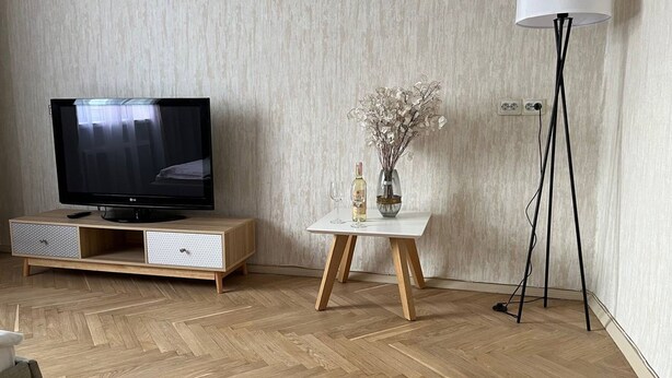 Un soggiorno moderno con una TV a schermo piatto su un mobile in legno, un piccolo tavolino con un vaso e una bottiglia di vino, e un'elegante lampada da terra.