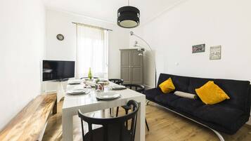 Un salon moderne avec un coin repas, comprenant un canapé avec des coussins jaunes, une table à manger en bois et un téléviseur fixé au mur.