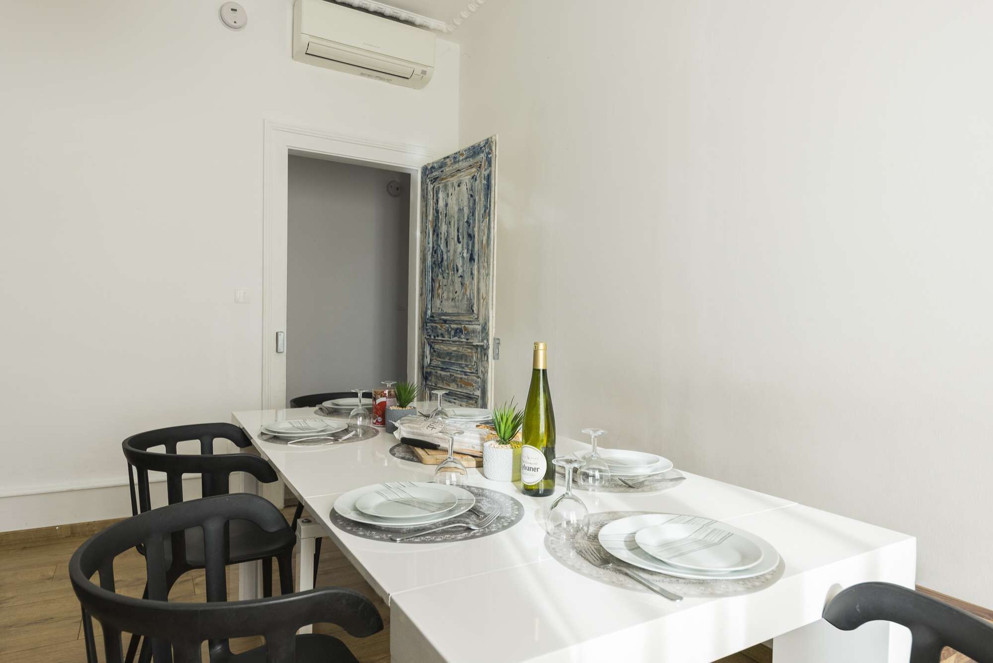 Un coin repas avec une table blanche dressée pour quatre personnes, comprenant des assiettes, des couverts et une bouteille de vin.