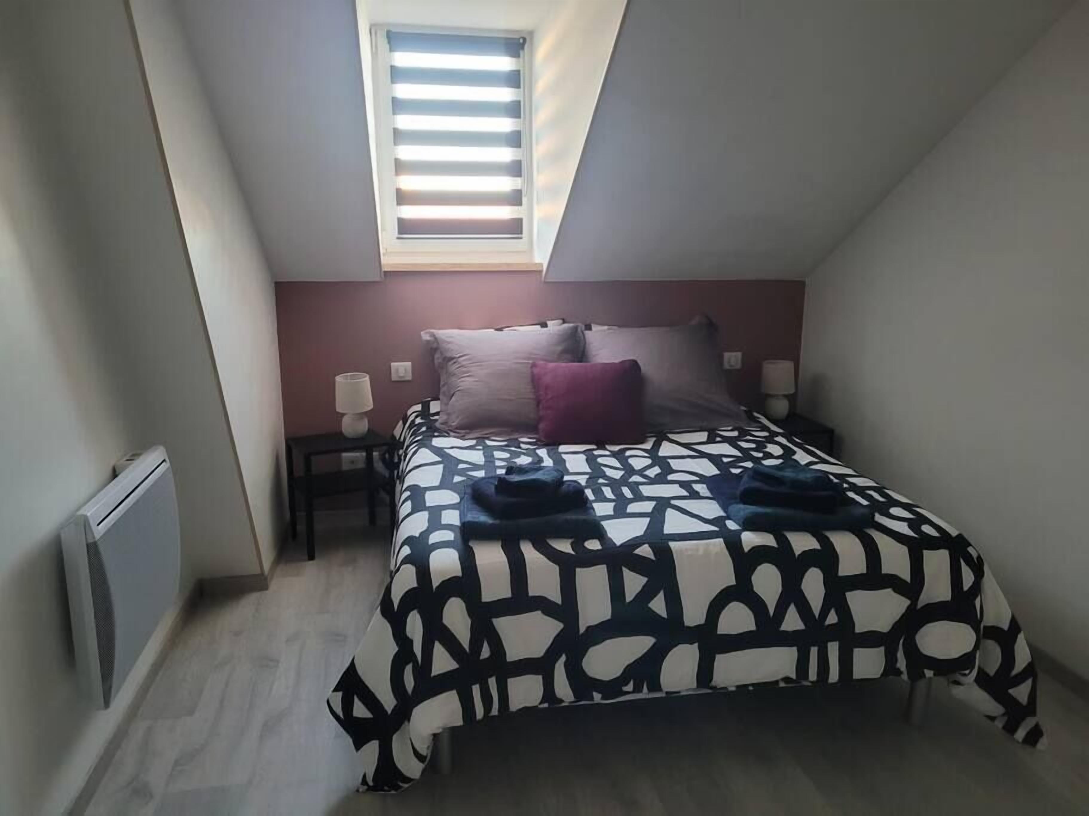 Een kleine, moderne slaapkamer met een schuine kap en dakraam, met een bed met een beddegoed met een geometrisch patroon, een nachtkastje en een wandradiator.