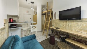 A compact living space with a kitchenette, a TV mounted on the wall, a blue sofa, and a patterned rug. - St Léon Strasbourg - Studio - 3 Pers (STRASBOURG)