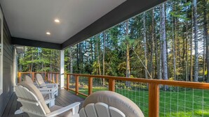 A covered deck with Adirondack chairs and a table, overlooking a forested area. - Brand New October 2023! Ultimate Golfers House in Bandon! Adjacent Sheep Ranch! (Bandon)