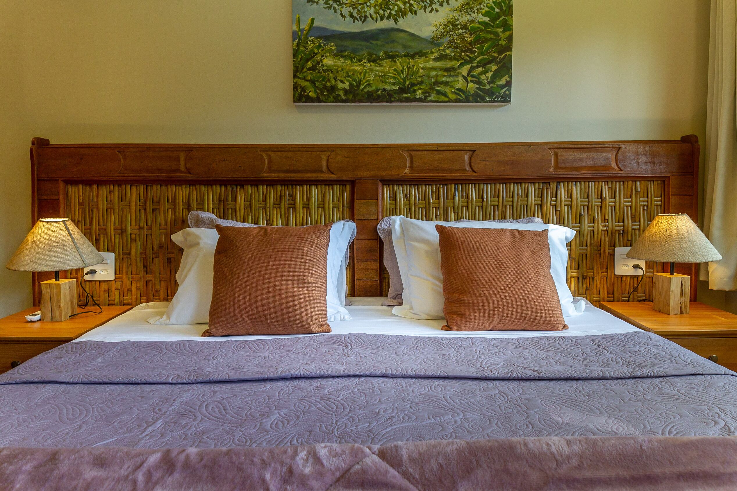 Uma cama de casal com cabeceira de madeira, duas mesas de cabeceira com luminárias e um quadro de uma paisagem na parede.