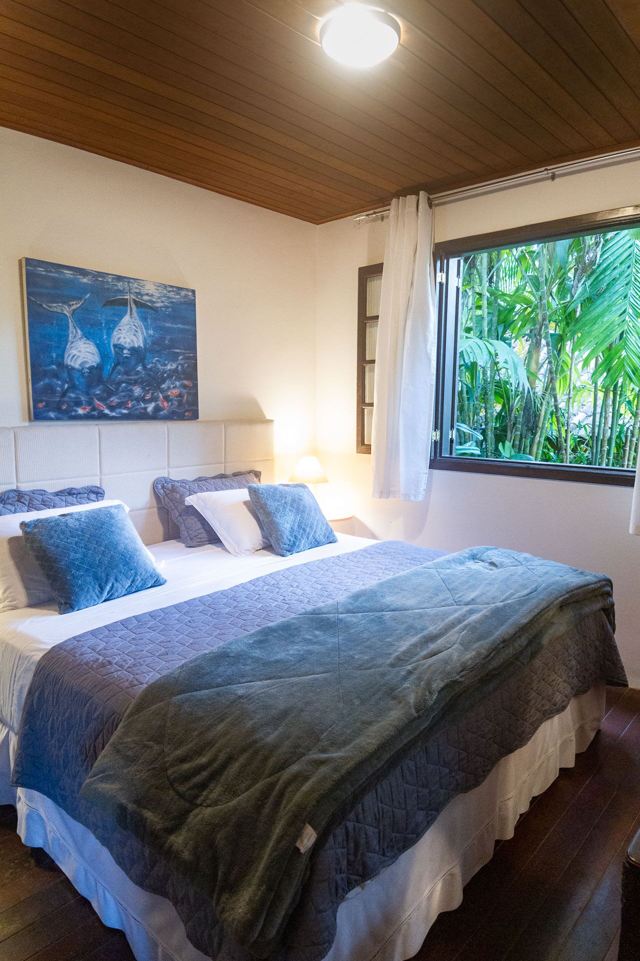 Quarto com cama, almofadas, um quadro, janela com cortinas e luminária no teto.