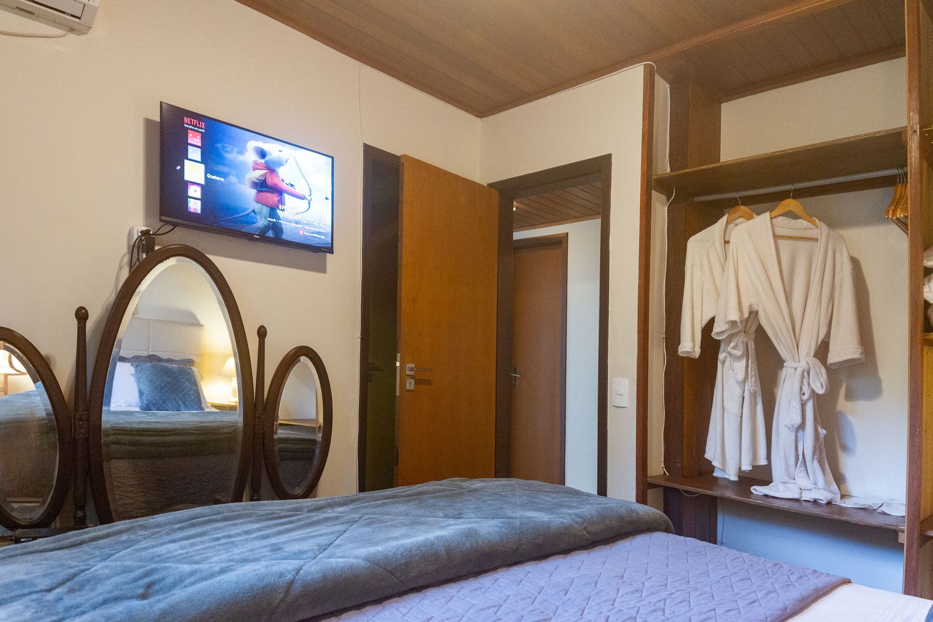 A hotel room with a bed, a TV mounted on the wall, a wooden wardrobe, and a mirror.