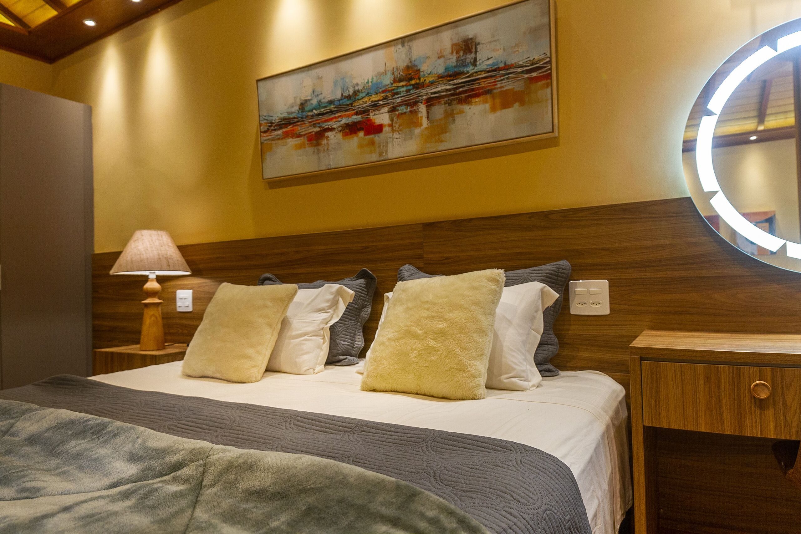 Un lit bien fait, avec une couette grise et blanche, un oreiller beige moelleux et un oreiller gris. On y voit une tête de lit en bois, une table de chevet avec une lampe et un miroir fixé au mur.