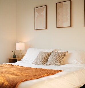 A neatly made bed with white linens and striped pillows, a bedside lamp, and two framed artworks on the wall.