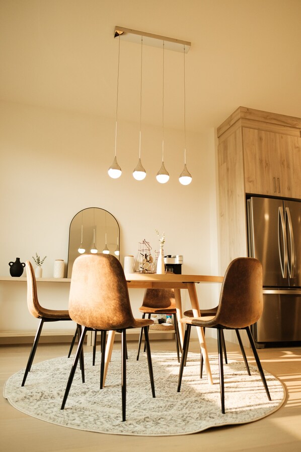 A modern kitchen with a wooden dining table, four chairs, a refrigerator, pendant lights, and a round rug.