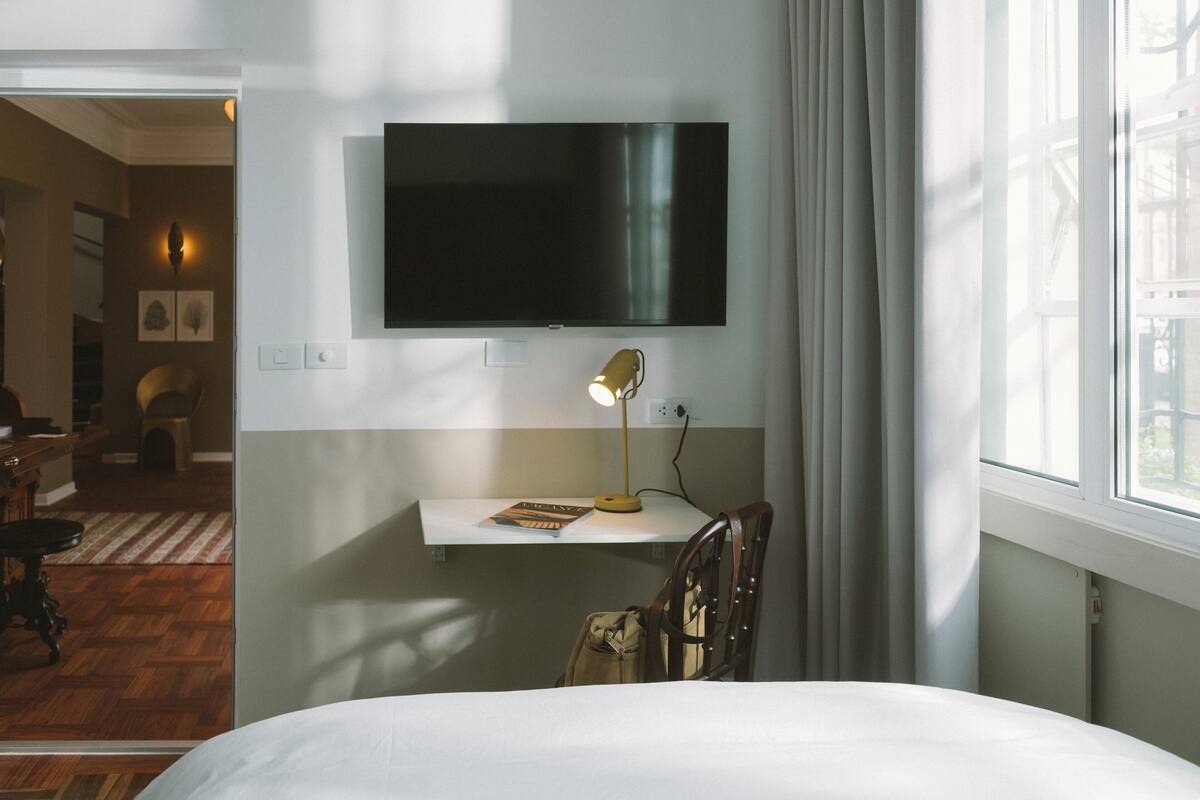 A hotel room with a bed, a desk with a lamp, a TV mounted on the wall, and a window with curtains.