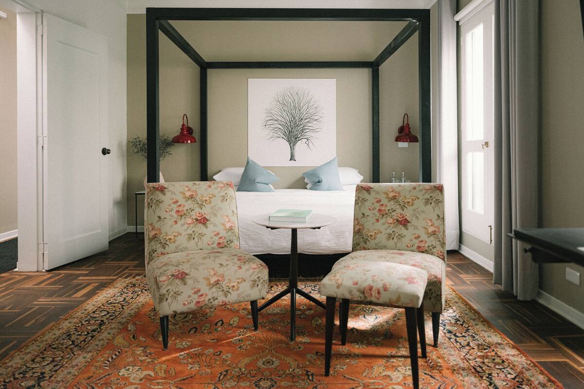 A bedroom with a four-poster bed, two floral armchairs, a small round table, and a large window with curtains.