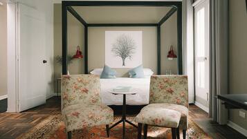 Ein Schlafzimmer mit einem Himmelbett, zwei floralen Sessel, einem kleinen runden Tisch und einem großen Fenster mit Vorhängen.
