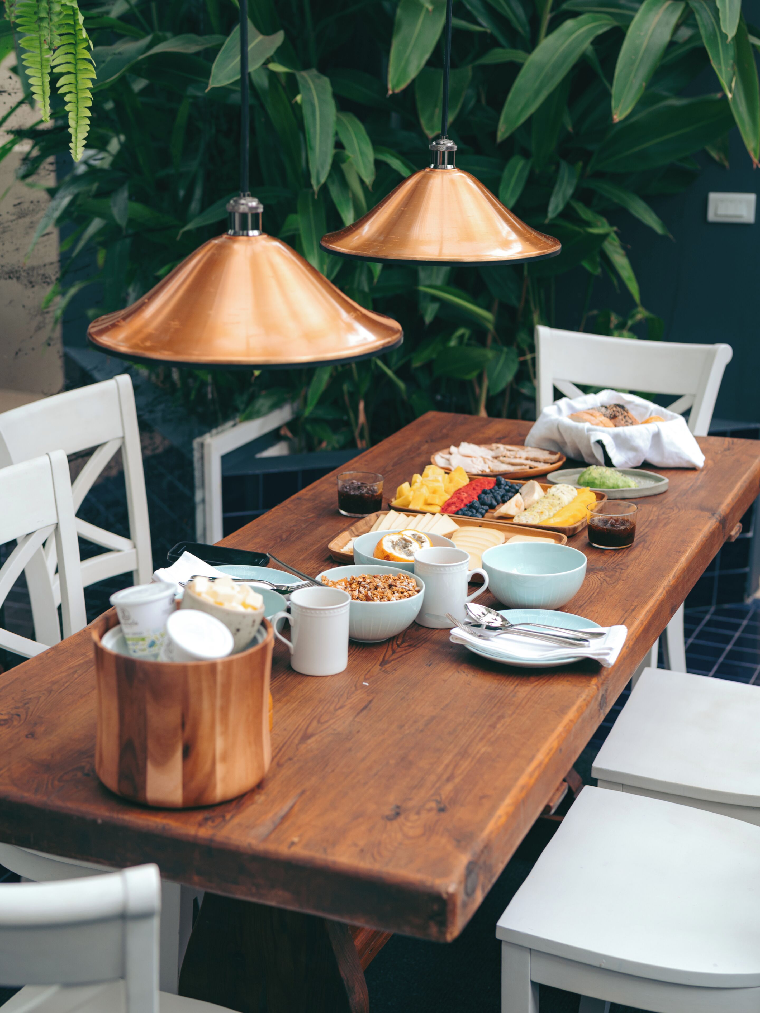 A wooden dining table with various food items and utensils, surrounded by white chairs, with hanging copper pendant lights and green foliage in the background.