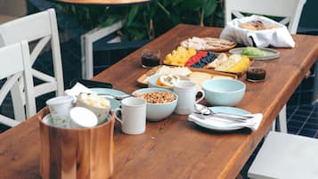 A wooden dining table with various food items and utensils, surrounded by white chairs, with hanging copper pendant lights and green foliage in the background.