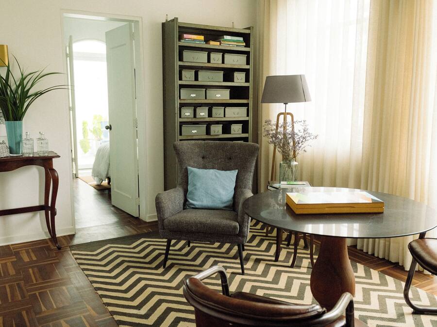 Ein Zimmer mit Holzboden, einem grauen Sessel mit blauem Kissen, einem runden Tisch mit Lampe, einem Bücherregal und einem Blumentopf.