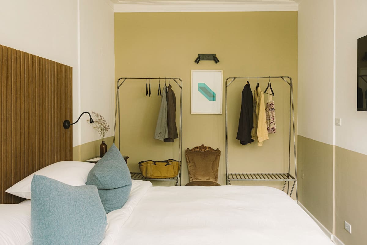 A hotel room with a bed, a wardrobe, a bedside table with a vase, and a hanging clothes rack.
