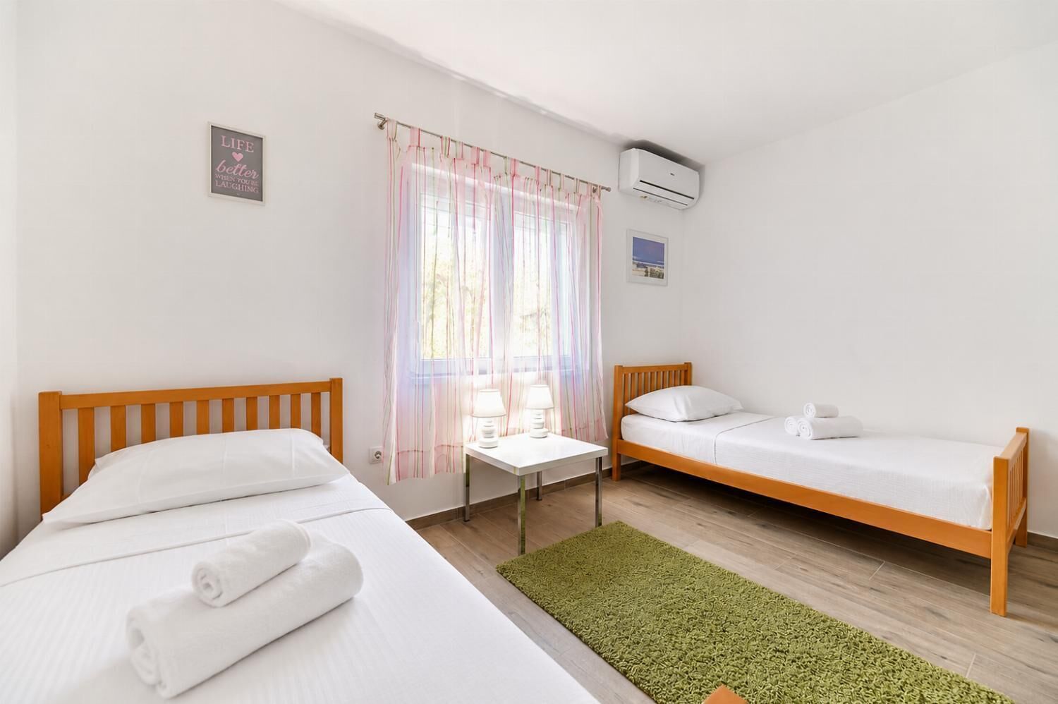 Una habitación con dos camas, una mesita pequeña, un tapete verde y un ventanal con cortinas de gasa.