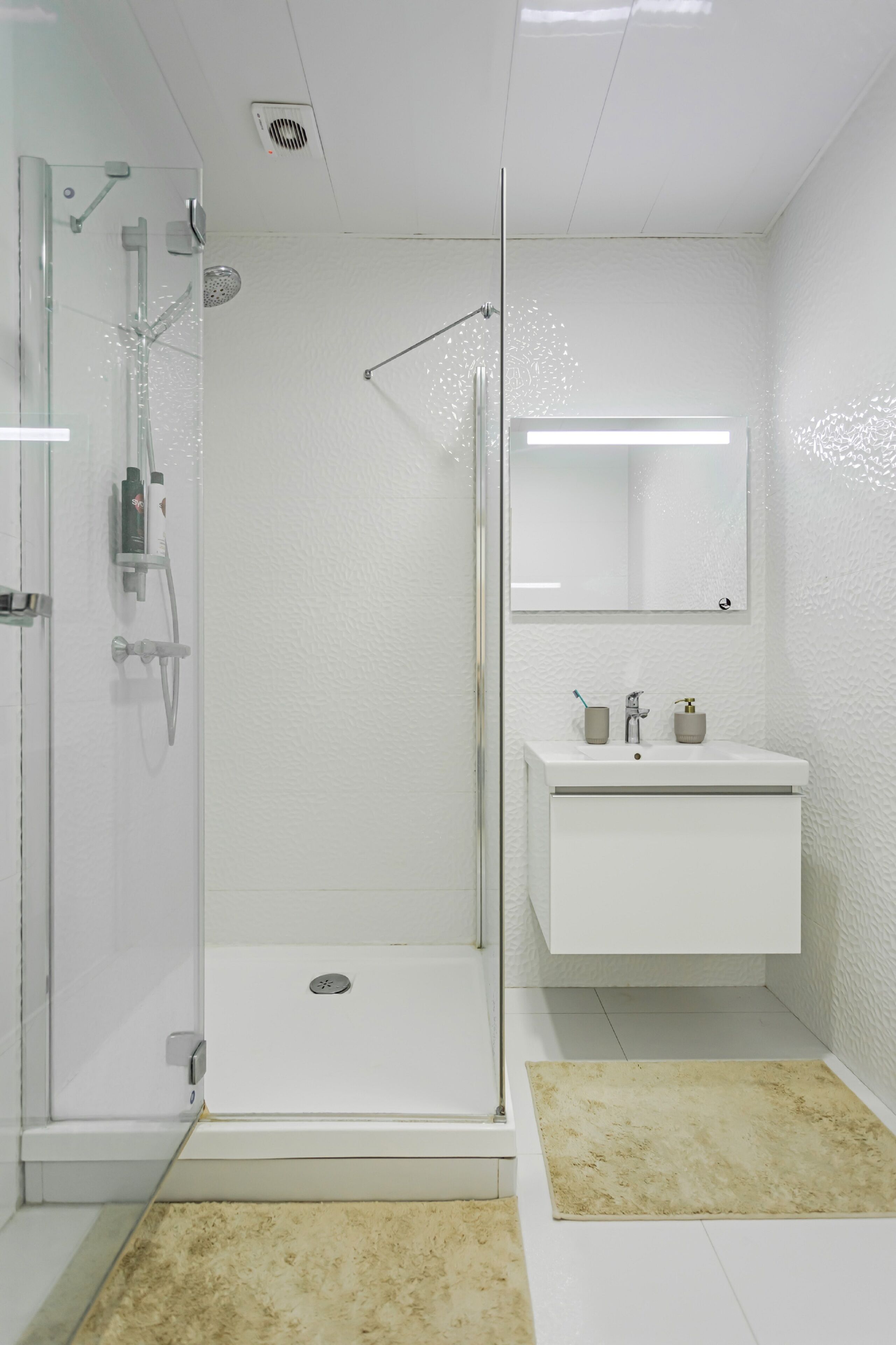 Ein modernes Badezimmer mit einer gläsernen Dusche, einem weißen Waschbecken mit Spiegel darüber und beigen Teppichen auf dem Boden.