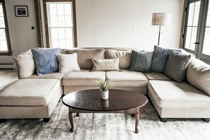 Ein Wohnzimmer mit einem beigefarbenen Ecksofa, einem runden Couchtisch mit Pflanze und einem großen Fenster, das natürliches Licht hereinlässt.