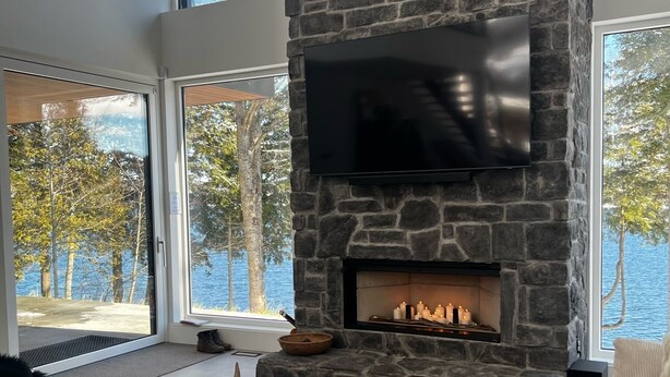 A modern living room with a stone fireplace, a flat-screen TV, and large windows overlooking a lake.