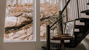 A spacious room with large windows offering a view of rocky terrain and trees. A wooden staircase with a black railing leads to an upper level.