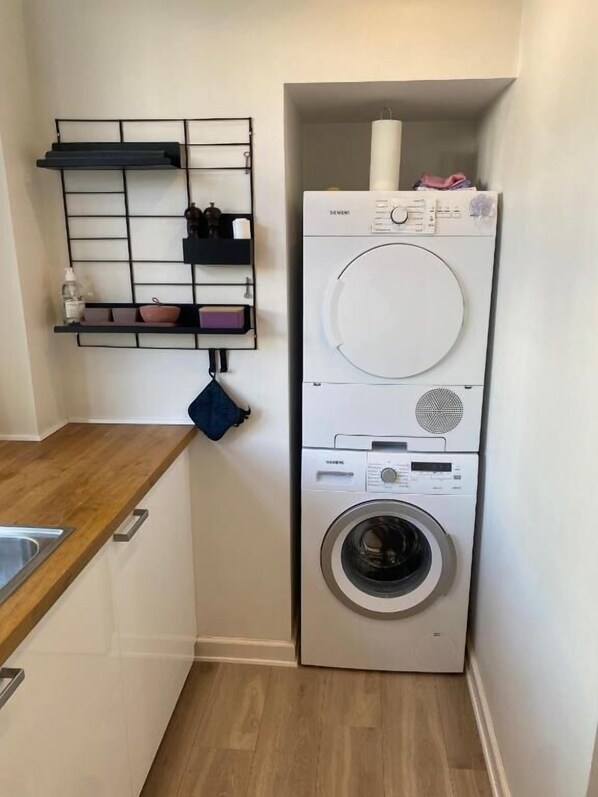 A compact laundry room with a built-in washer and dryer, a wooden countertop, and a metal shelf unit with various items. - Central Lyngby Apartments (Kongens Lyngby)