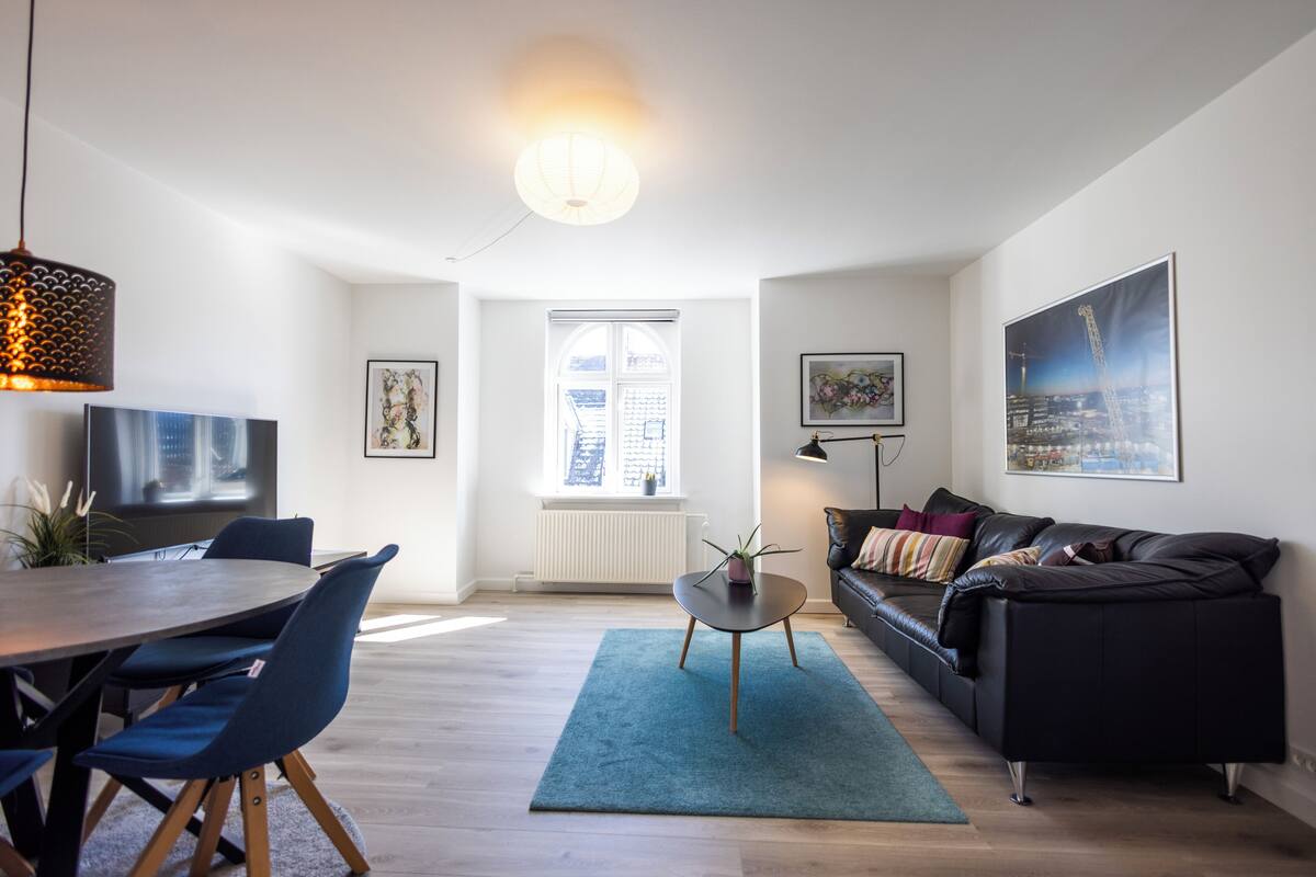 A modern living room with a dining area, a black leather sofa, a blue rug, and a large window with a view of the city.