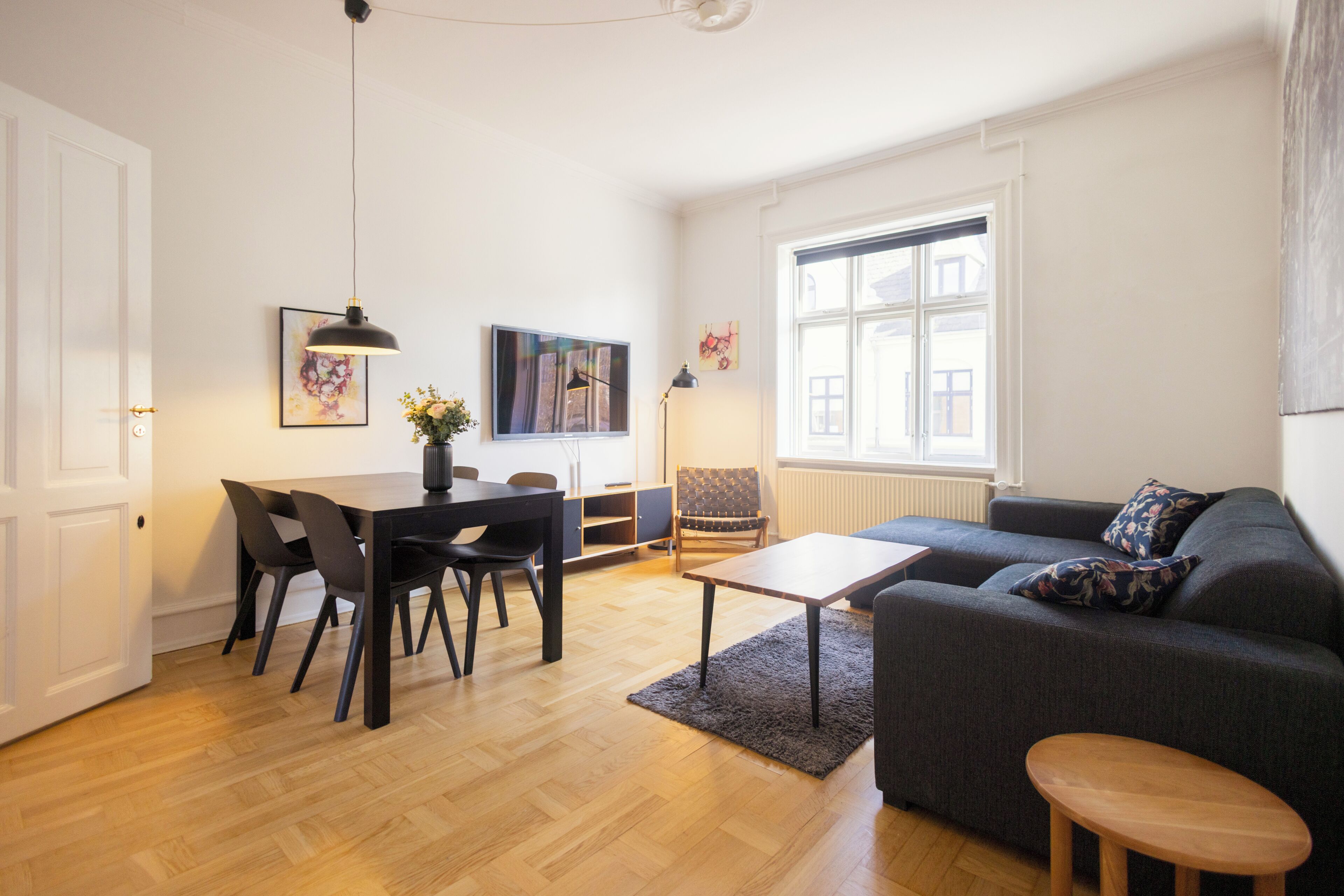A modern living room with a sofa, a dining table with chairs, a TV, and a window with blinds.