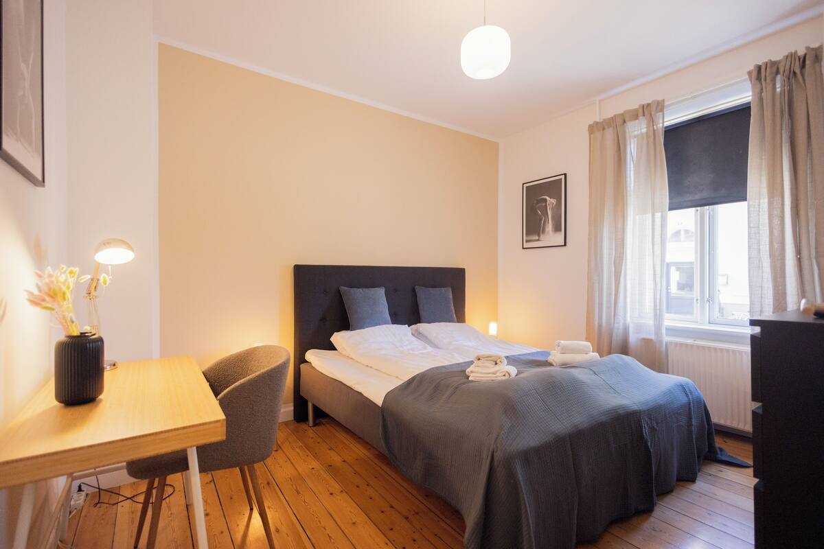 A hotel room with a bed, bedside table, chair, and a window with curtains.