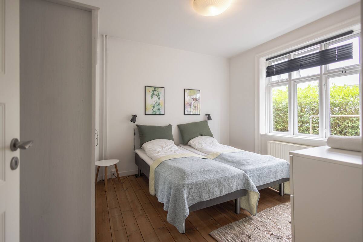 A bedroom with two beds, a small table, and a window with blinds.