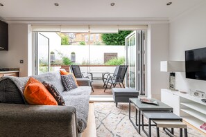 A modern living room with a sofa, coffee table, and TV, a balcony with chairs, and a city view.