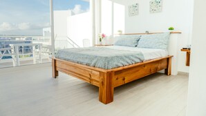 A modern bedroom with a wooden bed, large windows, and a balcony with a view of the sea.