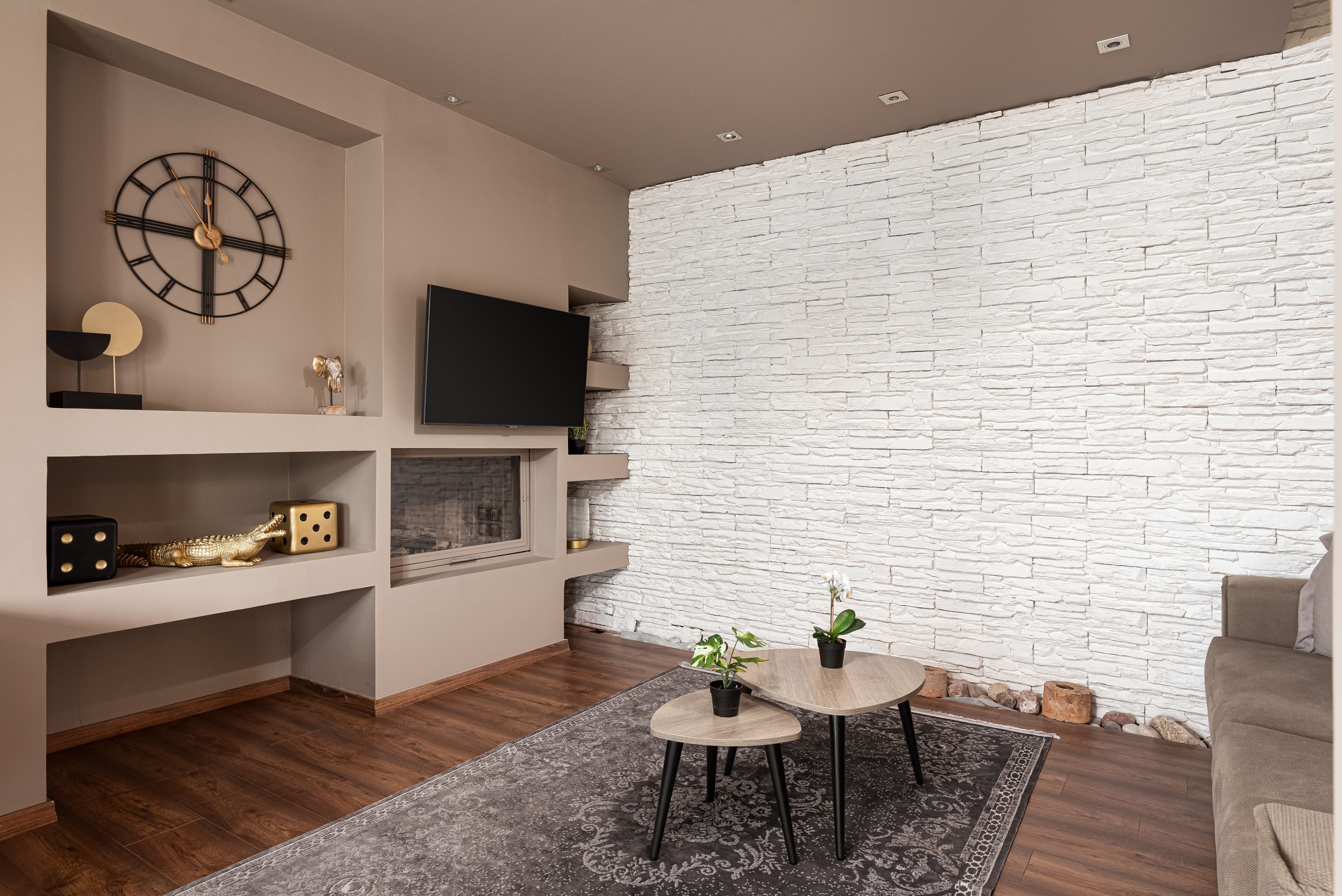 Una sala de estar moderna con una pared de piedra, un reloj de pared, un televisor de pantalla plana y una pequeña mesa redonda con dos plantas en macetas.