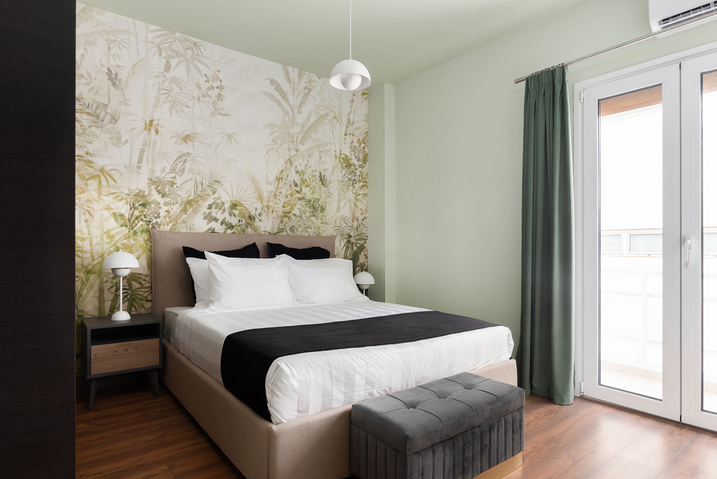 A bedroom with a bed, bedside tables, a lamp, a bench, a window with curtains, and a wall with a floral pattern.