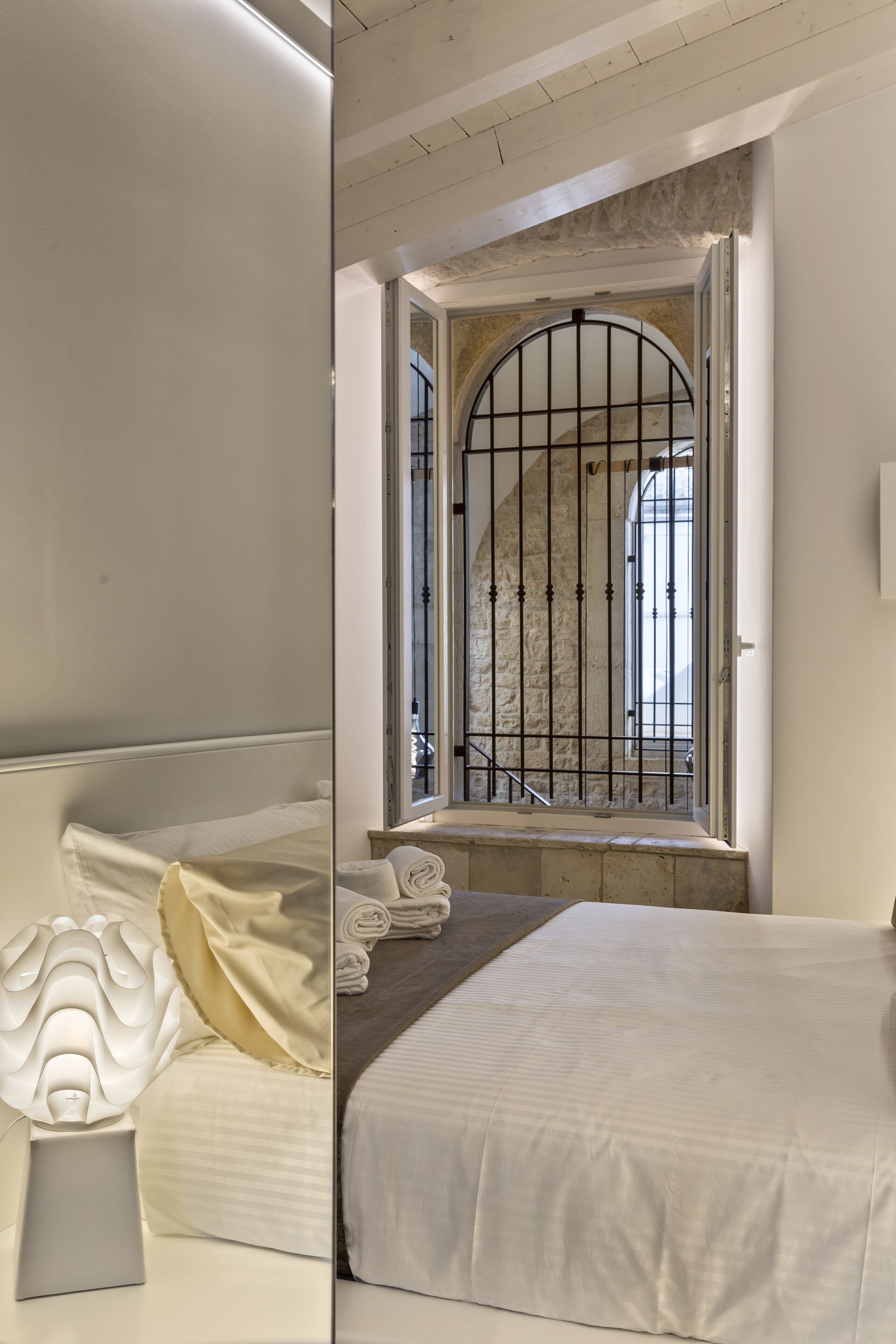 Une chambre à coucher avec un grand lit, une table de chevet avec une lampe et une fenêtre ornée d’un grillage métallique.