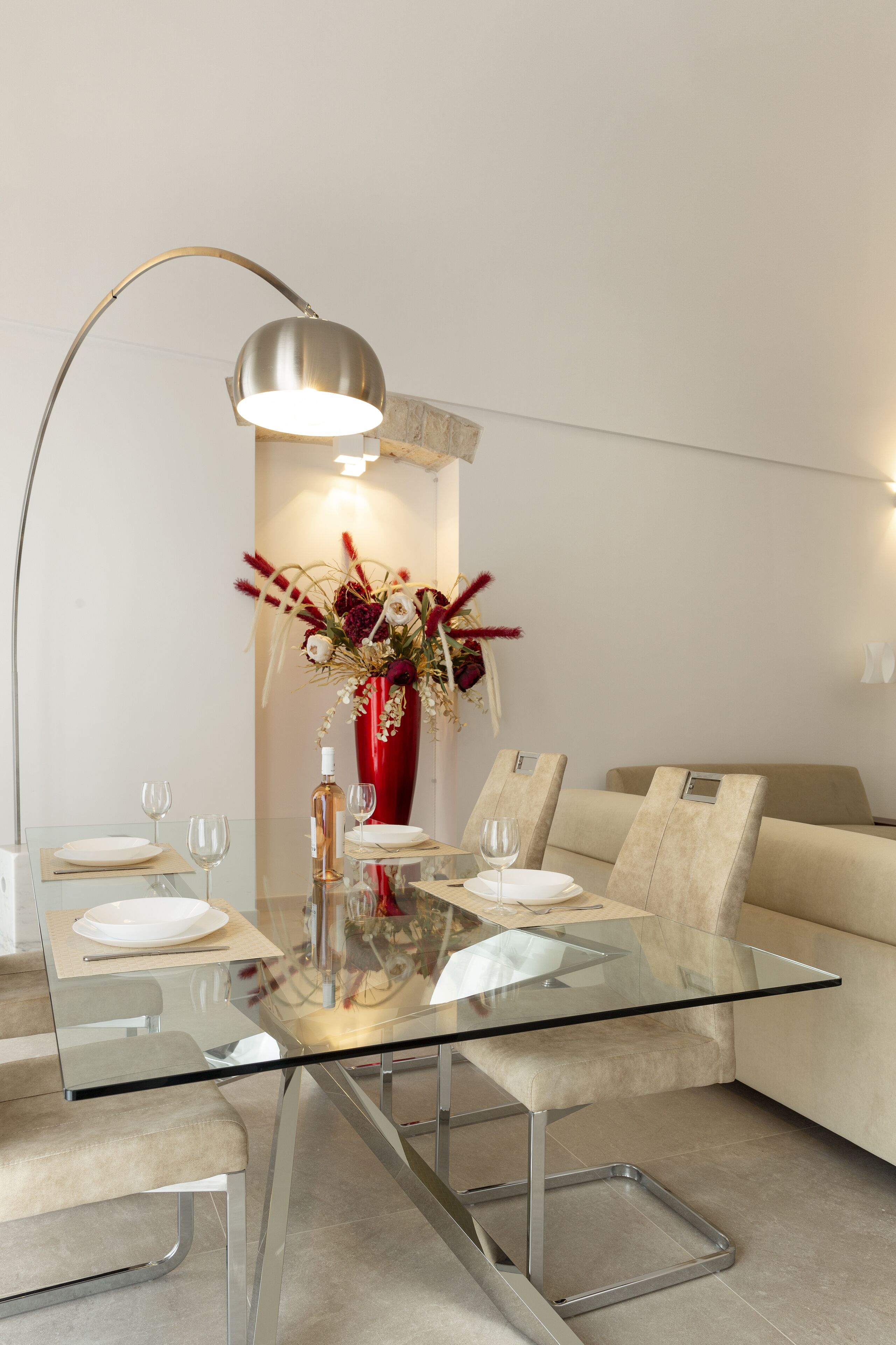 Un espace repas moderne avec une table en verre, deux chaises beiges, un vase rouge orné de fleurs et un lampadaire.