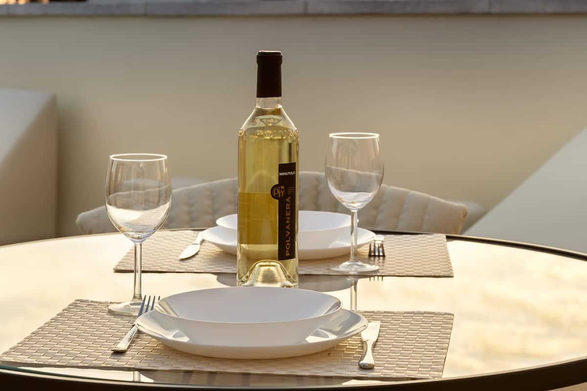 A table set for two with a bottle of white wine, two wine glasses, and plates, overlooking a historic building with a clock tower during sunset.