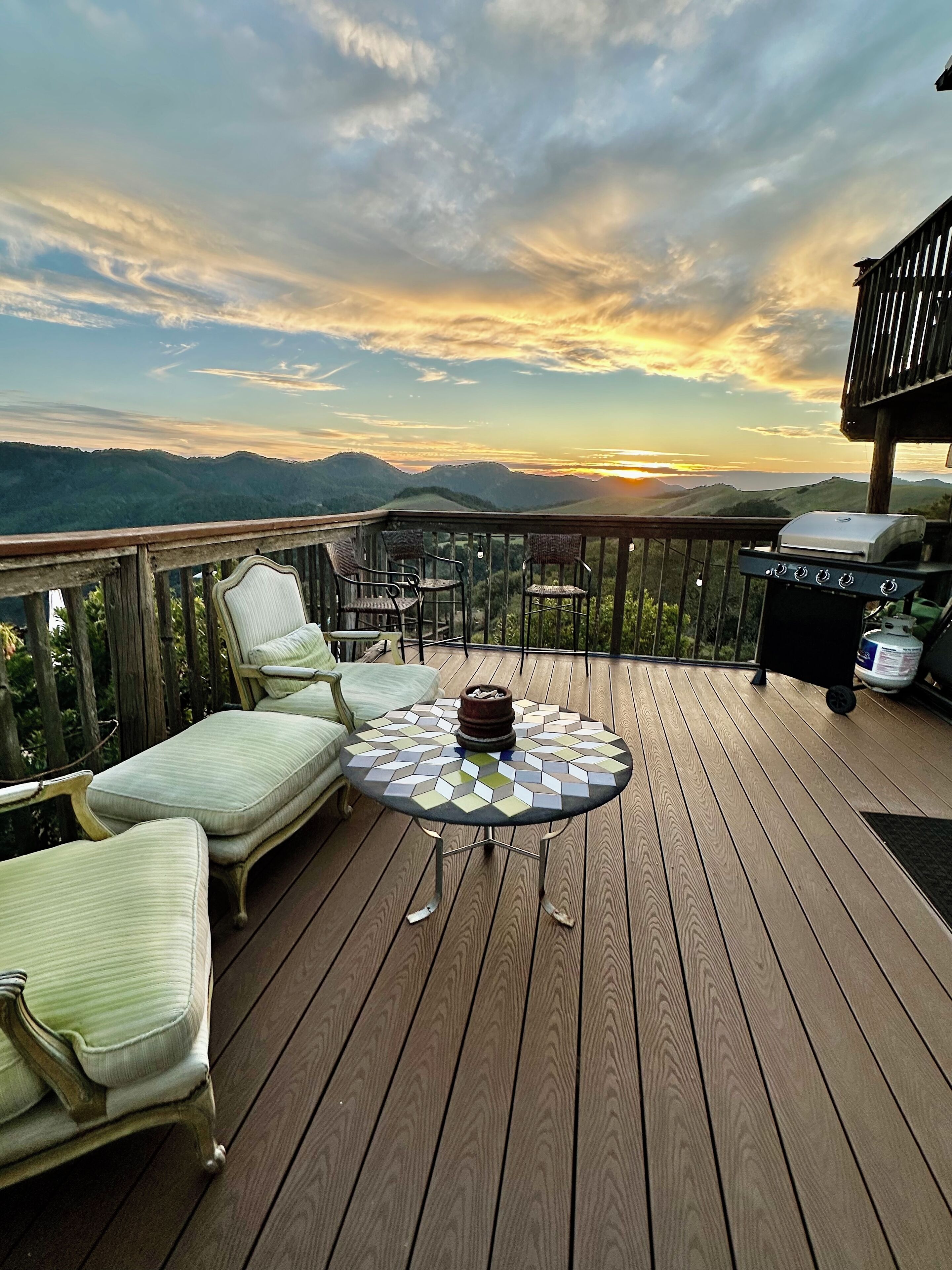 A wooden deck with a round table and chairs, a grill, and a view of the sunset over a mountain range.