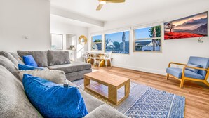 Een moderne woonkamer met een grijze bank, blauwe kussens, een houten salontafel en een blauw gestreept vloerkleed. Er is een eethoek met een witte tafel en stoelen, en een groot raam met uitzicht op een berglandschap.