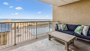 Un balcone con vista sulla spiaggia e sull'oceano, una piscina, un divano con cuscini e un tavolo di legno.