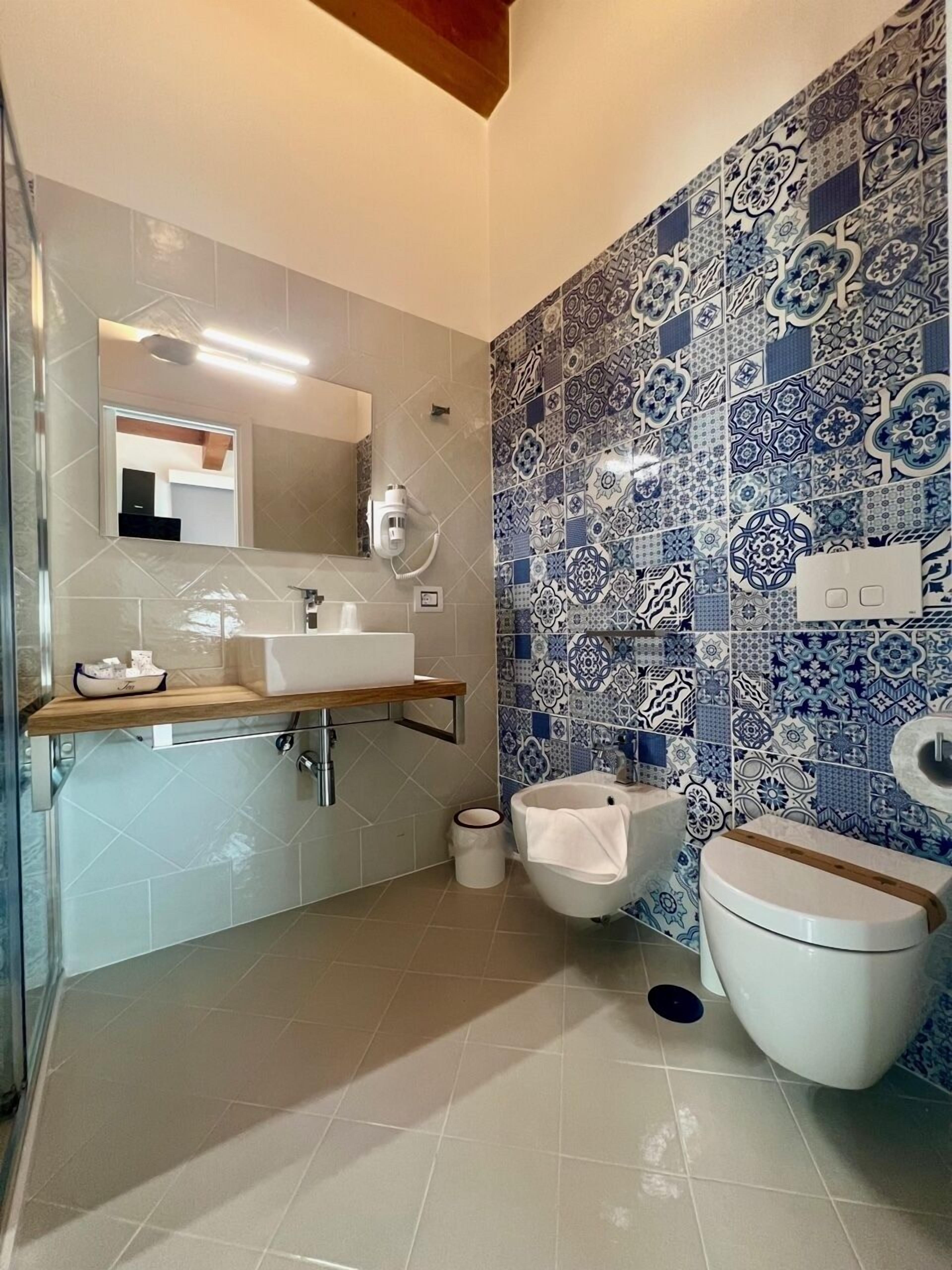 Un baño con piso de baldosas color beige, un lavamanos blanco con cubierta de madera, un inodoro blanco y una pared con baldosas azules y blancas.