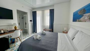 A hotel room with a bed, a desk with a lamp, a chair, a TV, and a painting of a coastal scene.