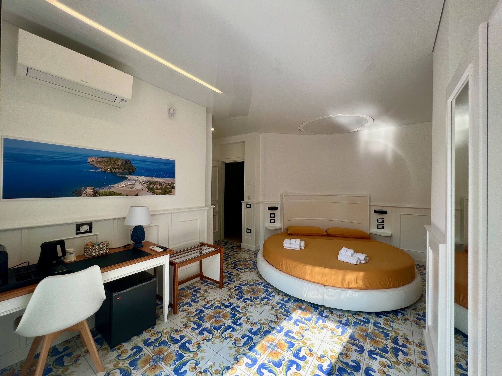Ein Hotelzimmer mit einem Bett, einem Schreibtisch, einem Sessel, einem kleinen Tisch, einer Lampe und einem Gemälde einer Küstenlandschaft.
