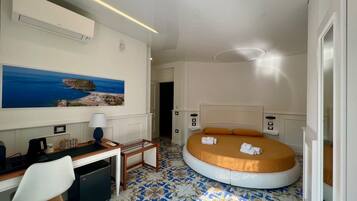 Ein Hotelzimmer mit einem Bett, einem Schreibtisch, einem Sessel, einem kleinen Tisch, einer Lampe und einem Gemälde einer Küstenlandschaft.